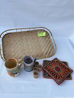 Photo showing woven wire bread basket, two ceramic mugs with abstract glaze, four small wooden cups, and two colorful floral woven coasters.