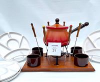 Full view of the vintage fondue set displaying the gradient fondue pot on metal stand with burner, six fondue forks with colored tips, four brown dip bowls, wooden serving tray, and two white divided ceramic plates.