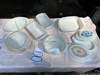 Full view of assorted Fire King and Pyrex bake and serving ware placed on a towel backlit outdoors showing divided dishes, rectangular loaf pans, casserole dishes and small serving dishes with floral decorations.