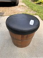 Front angle of barrel-style wooden stool with black cushioned seat on gravel and grass ground.