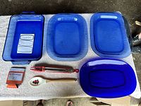 Top view of blue glass Pyrex lasagna pan, three blue platters, red tongs, wooden spoon, and Sharpie marker on white cloth