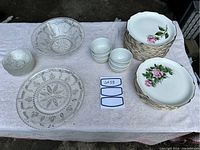 Photo showing 15 large rose patterned dinner plates stacked and 9 medium rose patterned plates stacked, 6 nappy dishes with scalloped edges, large and small pressed glass bowls, small white dessert bowls, and a rectangular serving platter.