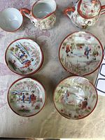 Top-down view of four hand-painted small plates and three porcelain vessels including bowl-shaped cup, creamer and sugar bowl with lid