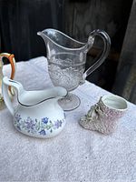 Clear pressed glass pitcher, porcelain creamer with purple floral pattern, and ceramic boot planter with floral decoration on white textured surface
