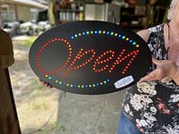 Front view of oval black metal sign with 'Open' spelled out in red LED lights, with additional blue and green LED accents in a semicircle pattern.