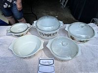 Five Pyrex dishes displayed on towel, including dishes with floral and vegetable patterns and clear glass lids.