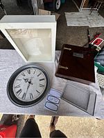 Photo showing a white shadow box frame, large round clock with Roman numerals, and a dark brown wooden keepsake box shaped like a book with metal clasps.