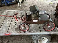 Side profile of the antique small wagon replica showing red wheels, brown body, and black cushioned seat.