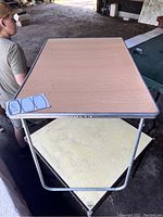 Rectangular folding table with light brown laminate top and gray trim, metal folding legs seen from an angle.