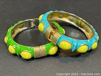 Two hinged enamel bangle bracelets lying overlapping on black surface showing green and blue colors with yellow dot accents