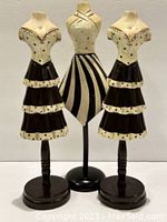 Front view of three painted wood dressmaking forms, two with ruffled skirts and one with striped skirt, standing side by side.