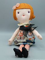 Full front view of the cloth doll sitting upright showing ginger bob hair with pink organza bow, sleeveless dress with large black bow, red and white diamond patterned legs, and black cloth shoes.