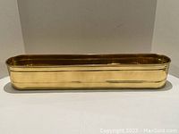 Wide angle front view of long narrow brass planter showing smooth shiny exterior with polished finish and rounded ends.