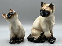 Pair of vintage ceramic Siamese cat figurines side by side, showcasing size difference and pose details.