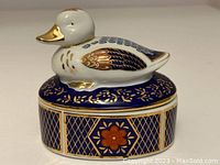 Front angle showing white porcelain duck figurine with gold beak, accented wings, sitting atop cobalt blue and gold decorated oval box base.