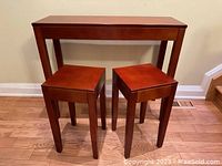 Photo showing front view of one tall rectangular red brown wood console table with two small square matching accent tables in front on a hardwood floor.