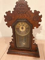 Front view of antique gingerbread wooden clock with visible carved details, Roman numeral dial, and glass door.