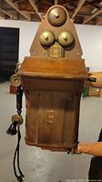 Front view of antique JYDSK wall telephone showing dual metal bells, emblem, hinged wooden front panel, and attached handset with frayed cord