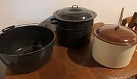 Three enamel canning pots displayed on a wooden surface; black large pot with handle, black pot with lid and insert, and brown/cream pot with lid.