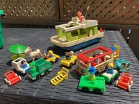 Full lot overview on table showing multiple vintage Fisher Price toys including Sesame Street sanitation truck, mini bus, plastic houseboat, yellow taxi with Sesame Street characters, small wooden peg figure, white cow, and various accessories.
