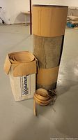 Two large rolls of wood veneer standing on a basement floor, one roll inside a cardboard box labeled Plywood Wrap.