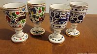 Set of four vintage hand painted porcelain wine goblets arranged in a row showing detailed colorful designs and gold rims.