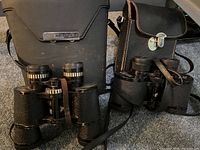 Two pairs of vintage binoculars with their respective carrying cases, displayed on a carpeted floor, showing overall condition and styling.