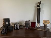 Wide shot of the lot showing all items on a wooden surface against a plain wall: Yashica-Mat camera with case, Kodak Brownie box camera, telephoto lens and case, tripod, and flash unit.