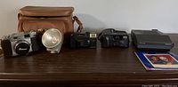 Full view of vintage cameras and accessories from left to right: Ricoh 300 camera with flash and brown case, Fuji camera, Yashica Mini camera, Polaroid Spectra 2 camera, Canon E08 camcorder and accessories