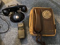 Lot showing all three telephones on carpet floor: brown rotary wall phone, black rotary desktop phone, and Motorola cell phone.