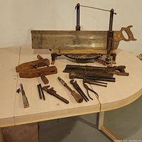 Photo showing a collection of vintage hand tools on a white table including a large Stanley miter saw, wooden hand planes, multi-bit screwdriver, wrenches, and other metal tools.