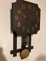 Front view of the dark wood square clock face with gold-tone numbers and matching hands. Shows carved corners in the clock face and the pendulum below.