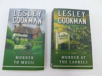 Two paperback books side by side: 'Murder to Music' with a country house cover and 'Murder at the Laurels' with a green cover and signpost.
