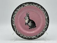 Full view of ceramic plate showing pink background with black and white bunny illustration and laurel leaf border.