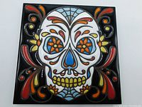 Front view of the hand-glazed Day of the Dead skull ceramic tile showing the colorful raised relief design with floral eyes and ornamental accents on black background.