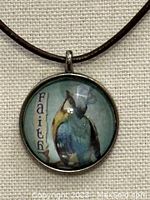 Close-up of round glass pendant showing the word 'FAITH' and a blue crowned bird image inside a metal frame.