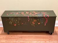 Front view of the olive green wooden chest with floral handpainted design and caster wheels.