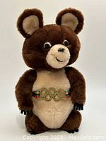 Full view of the brown and beige Misha bear plush toy standing upright showing its facial expression and belt with Olympic rings.