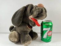 Front angle showing gray plush dog with large floppy ears, brown nose, red tongue, and beige paw pads. 7Up soda can for scale.