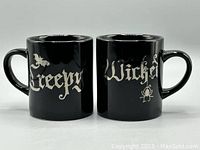 Front view of two black ceramic mugs showing 'Creepy' on one with bat, 'Wicked' on the other with spider.