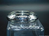 Polished hammered metal silver-tone bangle placed on clear block showing curved hammered surface detail.