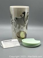 Front view of Starbucks ceramic tumbler showing iridescent silver siren design and included light green plastic lid, with informational materials.