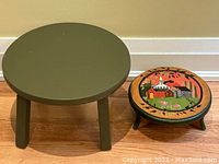 Two round wooden stands; the smaller one decorated with a handpainted colorful village scene on top, the larger one plain olive green.