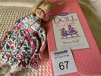 Goebel Doll Club Bette Ball Doll laying on beige knit surface wearing floral dress and crochet vest alongside original pink Doll Club box