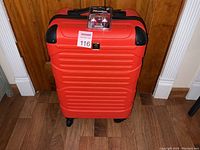 Front view of the red hard shell suitcase with black corner protectors and wheels on the floor in front of a wooden door.