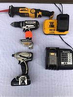 Five battery operated power tools and chargers on table: DeWalt reciprocating saw, Makita drill and impact driver, DeWalt and Makita chargers; no batteries present.