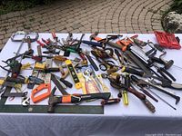 Full overview of assorted hand tools on a white tablecloth, arranged loosely showing variety including saws, clamps, hammers, pliers, screwdrivers, levels, and wrenches.