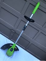 Full side view of green and black Greenworks electric grass trimmer showing handle, shaft, and cutting head with guard.