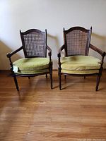 Front view of the two vintage cane backed chairs with pea green cushions on a hardwood floor against a plain wall.