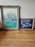 Full view of two framed artworks on wooden floor against beige wall - large floral painting by Mary Glasson and smaller dancer & swan print.